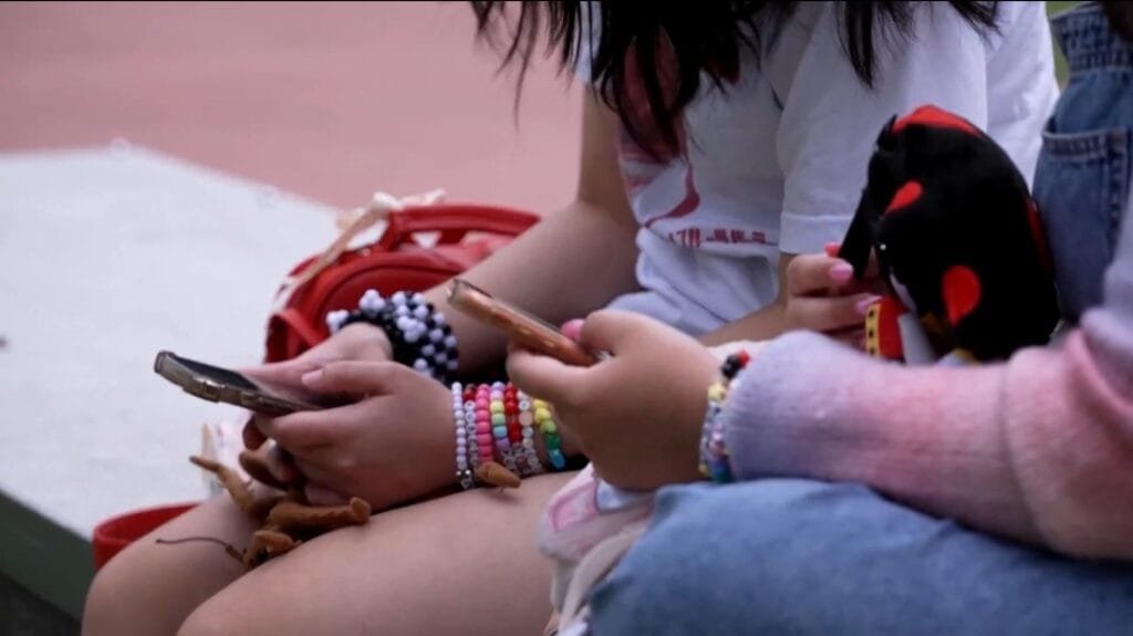 Two young people with colorful bracelets are engrossed in their phones. This image illustrates the potential impact of Australia's ban on social media for those under 16.