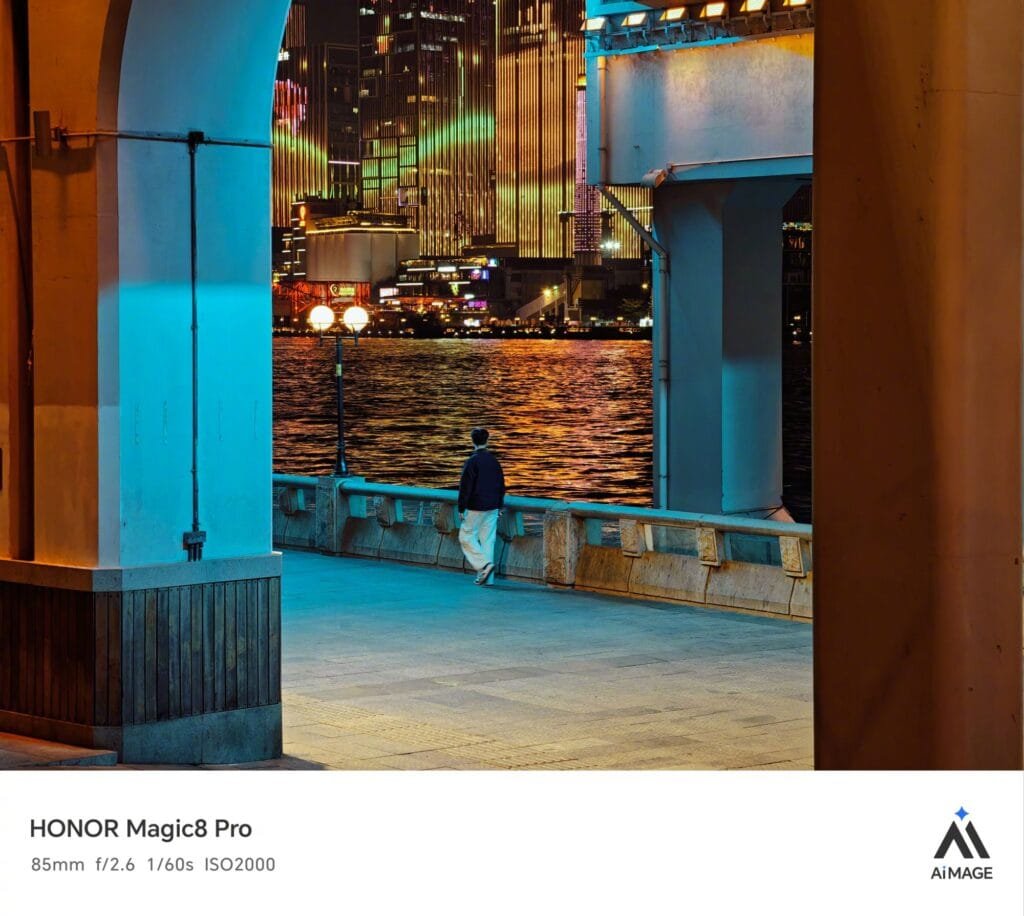 A nighttime city waterfront photograph taken with HONOR Magic8 Pro (85mm, f/2.6, 1/60s, ISO2000). The scene is framed by large architectural pillars (one lit blue, the other warm-toned), showing a person in dark top and light pants walking along a stone railing. Across the water, brightly lit skyscrapers reflect colorful lights on the rippling surface. The image includes HONOR Magic8 Pro branding and the "AIMAGE" logo at the bottom.