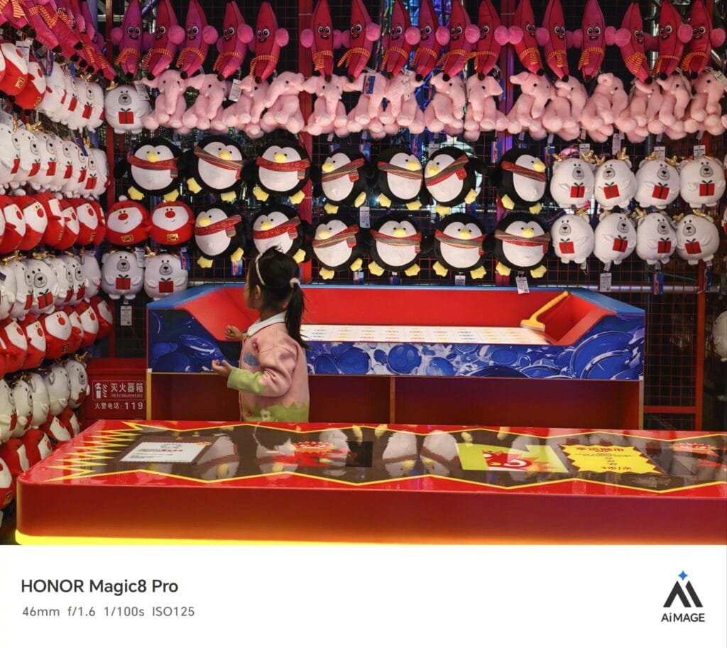 A photograph captured with HONOR Magic8 Pro (46mm, f/1.6, 1/100s, ISO125) shows a young child in a pink outfit standing at a vibrant carnival game booth. The background wall is densely lined with rows of plush toys—including red octopuses, pink bunnies, penguins, and other characters. A red-and-blue game counter sits in front of the child, with a "Fire Extinguisher Box" sign visible to the left. The image features HONOR Magic8 Pro branding and the "AIMAGE" logo at the bottom.