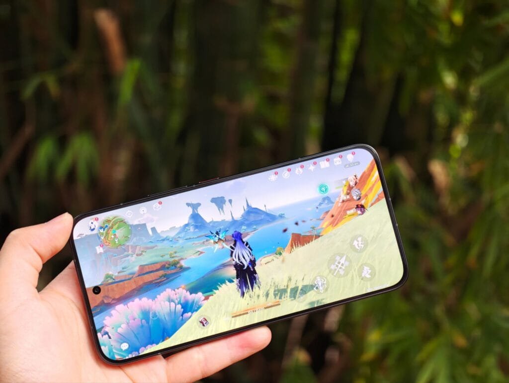 Redmi Turbo 5 Max to Debut with Dimensity 9500s | Gaming Test Performance 1 Redmi Turbo 5 Max smartphone being held in one hand, displaying a vibrant open-world game (likely Genshin Impact) on its 1.5K flat display. The device is positioned as a 2026 mid-to-high-end model, featuring flagship-level specs like MediaTek Dimensity 9500S processor, large-capacity battery, metal frame, and glass back, with a focus on smooth gaming performance. Background shows blurred green foliage, emphasizing the phone’s display clarity even in outdoor settings.