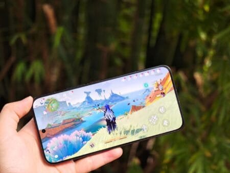 Redmi Turbo 5 Max to Debut with Dimensity 9500s | Gaming Test Performance Redmi Turbo 5 Max smartphone being held in one hand, displaying a vibrant open-world game (likely Genshin Impact) on its 1.5K flat display. The device is positioned as a 2026 mid-to-high-end model, featuring flagship-level specs like MediaTek Dimensity 9500S processor, large-capacity battery, metal frame, and glass back, with a focus on smooth gaming performance. Background shows blurred green foliage, emphasizing the phone’s display clarity even in outdoor settings.