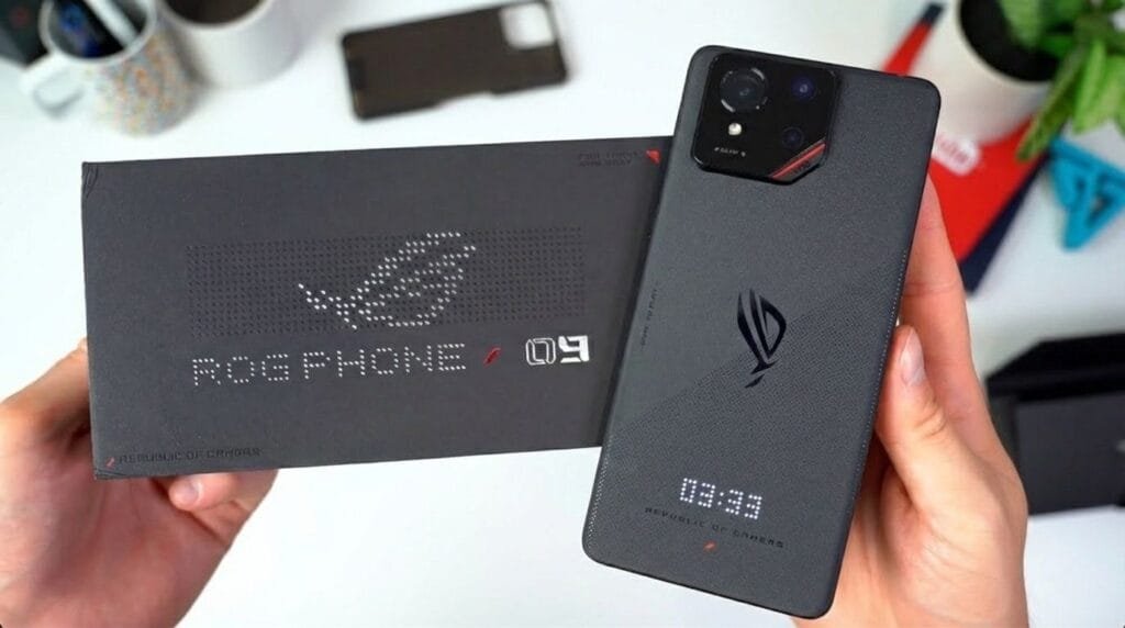 ASUS temporarily halts new phone launches in pivot to ‘Physical AI’ and commercial PCs Two hands holding an ASUS ROG Phone 9 and its black packaging box on a white surface. The phone’s back features the ROG logo, a rectangular camera module, and a small LED display showing '13:35'. The box has 'ROG PHONE' and '8' printed on it, along with the Republic of Gamers branding. Office items like a plant and containers are blurred in the background.