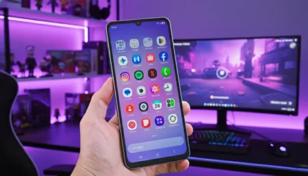 Samsung Galaxy A07 Launched in Philippines with 6 Years of OS Upgrades Hand holding a Samsung Galaxy A07 5G smartphone, displaying its home screen with various app icons. The phone is showcased against a purple-lit gaming setup background featuring a monitor, keyboard, and shelves with collectibles.