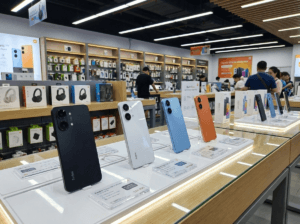 17697360485872214616227105110378 1 Redmi Turbo 5 and Redmi Turbo 5 Max smartphones displayed on a Xiaomi retail store demo table, showing multiple color options with price tags and accessory shelves in the background.