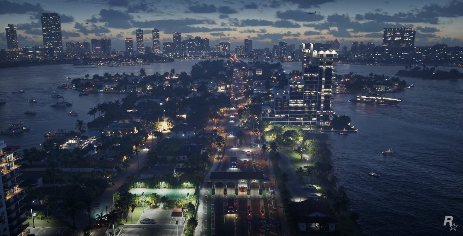 Dusk aerial view of a fictional tropical waterfront city (likely from Grand Theft Auto VI, indicated by the Rockstar Games logo), featuring a narrow island with a lit main road, residential homes, palm trees, a basketball court, and modern high-rises. Across the water, a dense city skyline glows under a cloudy twilight sky, with boats dotting the dark water.