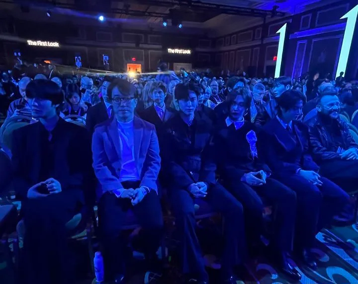 Attendees seated in rows at Samsung's First Look event during CES 2026. The venue is lit with blue lighting, with 'The First Look' signage visible in the background. A diverse crowd fills the space, and some individuals in the front row are dressed in formal dark attire, with one person wearing a light purple blazer. Large illuminated arrow decorations are present on the right, and the patterned carpet adds to the event setting.