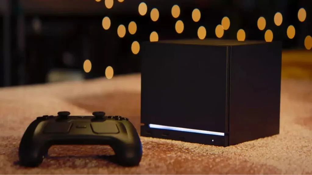 Black Steam Machine gaming console with a horizontal blue light strip at the base, placed next to a matching black wireless controller. Both rest on a textured light brown surface, with out-of-focus warm yellow bokeh lights in the background.