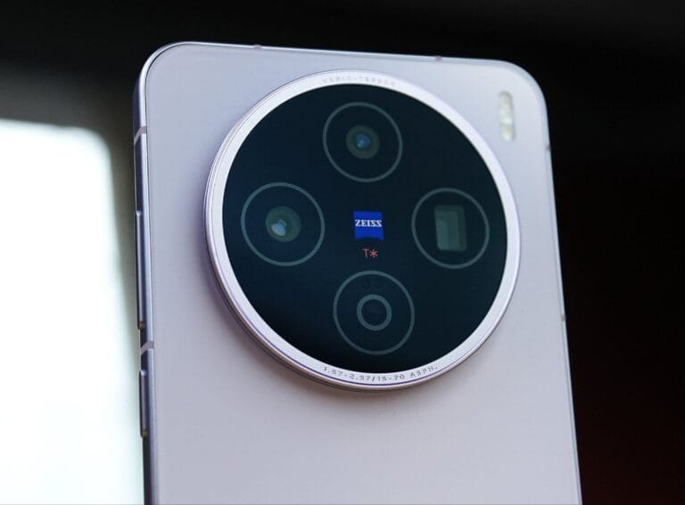 Vivo X200T Launched in India with Dimensity 9400+ and Packing a 6,200mAh Battery Close-up shot of the back of a light-colored Vivo X200T smartphone, highlighting its large circular camera module with a metallic ring. The module houses four lenses and features a blue Zeiss logo with a red "T*" marking at its center. Text indicating lens specifications is visible around the inner edge of the ring.