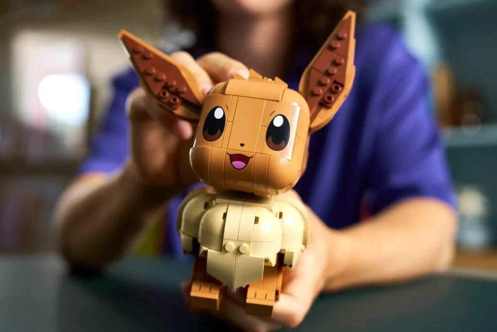 A detailed LEGO model of Pokémon character Eevee, featuring a brown head and limbs, lighter cream-colored chest, large pointed ears with visible brick details, big expressive eyes, and an open mouth showing a pink tongue. The model is being held up by a person wearing a purple shirt, with a blurred indoor background.