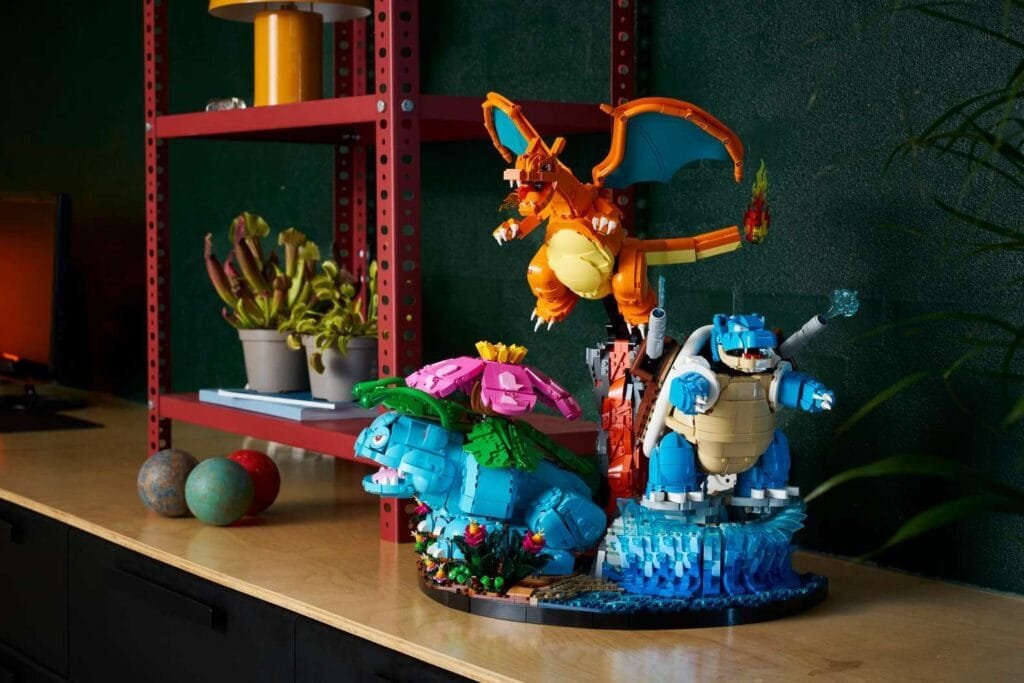 A detailed LEGO display featuring the final evolved forms of the Kanto starter Pokémon: Venusaur (blue body with green foliage and a large pink flower on its back, set among small plants), Charizard (orange with blue wings, posed mid-flight with a flame at the tip of its tail, perched above), and Blastoise (blue and beige shelled form, with water cannons firing, positioned on a textured blue water base). The set is placed on a light wood surface, with a red metal shelving unit in the background holding potted plants, a yellow lamp, and decorative spheres; the backdrop includes a dark green wall and hints of green foliage.