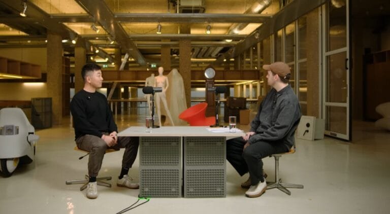 Carl Pei (right, brown cap) in industrial-style studio, seated opposite guest at mic-equipped table, discussing Nothing’s 2026 strategy; design mannequin/decor in background.