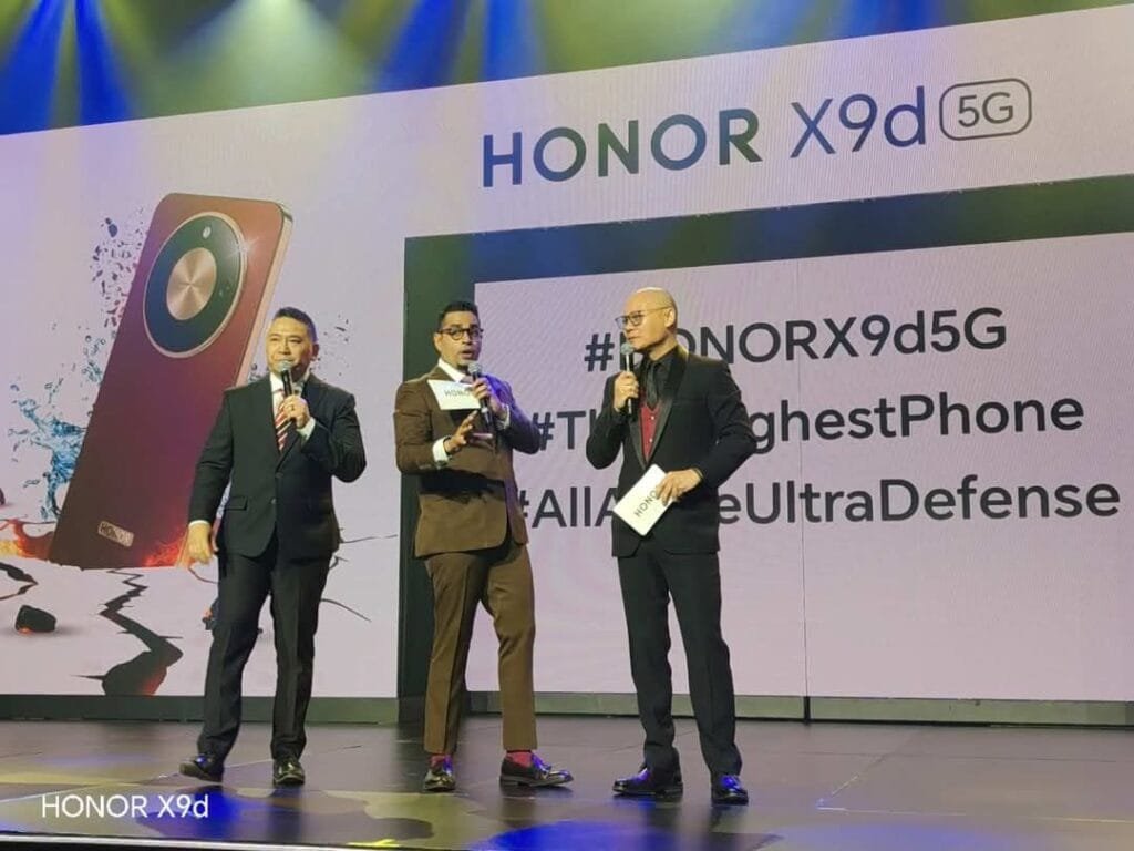 HONOR X9d 5G Launches in Philippines: 8,300mAh Battery, IP69K Protection, Tesla Cybertruck Raffle 1 Stage scene from the HONOR X9d 5G launch event. Three men in formal attire stand on stage holding microphones. A large backdrop displays 'HONOR X9d 5G' at the top, alongside a graphic of the Reddish Brown phone variant with visuals emphasizing water and impact resistance. Partial hashtags and feature-related text are visible on the screen, with stage lighting in blue and green tones. 'HONOR X9d' is labeled at the bottom left.
