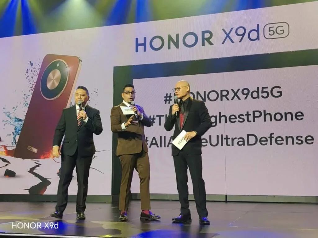 HONOR X9d 5G Launches in Philippines: 8,300mAh Battery, IP69K Protection, Tesla Cybertruck Raffle 1 Stage scene from the HONOR X9d 5G launch event. Three men in formal attire stand on stage holding microphones. A large backdrop displays 'HONOR X9d 5G' at the top, alongside a graphic of the Reddish Brown phone variant with visuals emphasizing water and impact resistance. Partial hashtags and feature-related text are visible on the screen, with stage lighting in blue and green tones. 'HONOR X9d' is labeled at the bottom left.