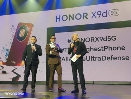 Stage scene from the HONOR X9d 5G launch event. Three men in formal attire stand on stage holding microphones. A large backdrop displays 'HONOR X9d 5G' at the top, alongside a graphic of the Reddish Brown phone variant with visuals emphasizing water and impact resistance. Partial hashtags and feature-related text are visible on the screen, with stage lighting in blue and green tones. 'HONOR X9d' is labeled at the bottom left.