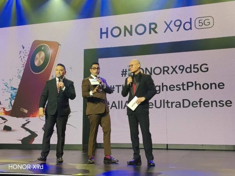HONOR X9d 5G Launches in Philippines: 8,300mAh Battery, IP69K Protection, Tesla Cybertruck Raffle Stage scene from the HONOR X9d 5G launch event. Three men in formal attire stand on stage holding microphones. A large backdrop displays 'HONOR X9d 5G' at the top, alongside a graphic of the Reddish Brown phone variant with visuals emphasizing water and impact resistance. Partial hashtags and feature-related text are visible on the screen, with stage lighting in blue and green tones. 'HONOR X9d' is labeled at the bottom left.