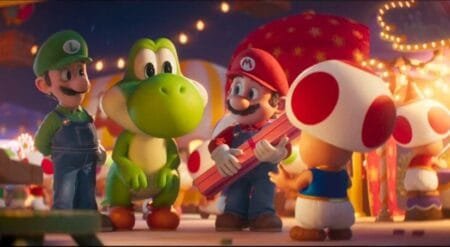 Mario holds a pink gift box while talking with Toad at a festive outdoor fair, as Luigi and Yoshi stand nearby under warm string lights in the Super Mario Galaxy movie.