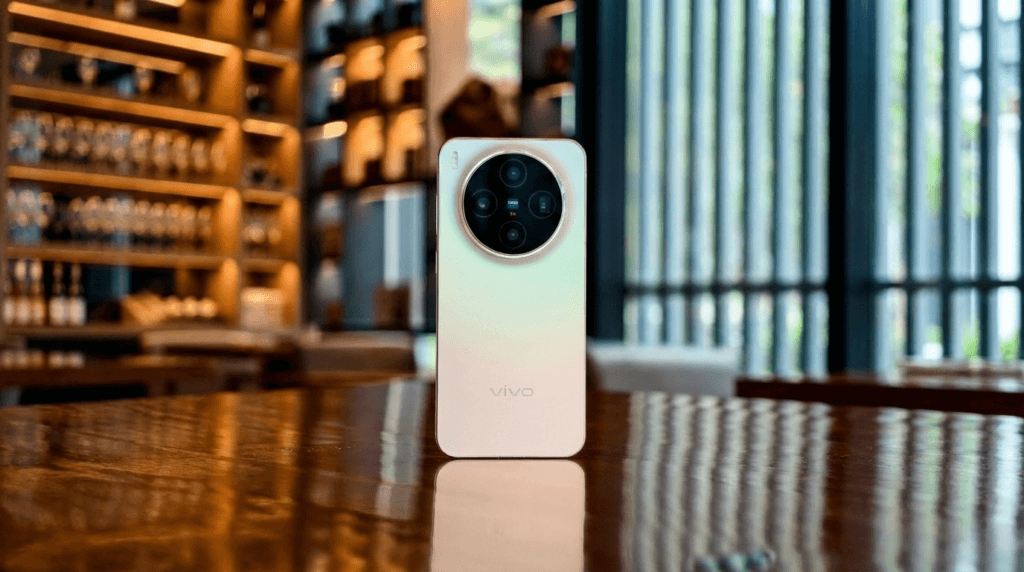 A rear view of a Vivo smartphone with a prominent, large circular ZEISS camera module, standing upright on a highly reflective wooden table. The back panel has a white to light gold gradient finish. The phone is reflected sharply on the table's surface. The background is a warmly lit, blurred interior, possibly an upscale bar or lounge, featuring shelving with glassware and vertical slats.
