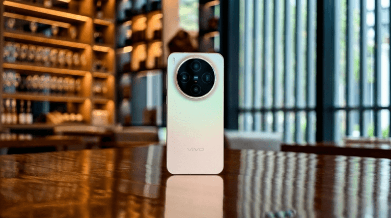 Vivo X300 Max Tipped to Be Powered by MediaTek Dimensity 9500 A rear view of a Vivo smartphone with a prominent, large circular ZEISS camera module, standing upright on a highly reflective wooden table. The back panel has a white to light gold gradient finish. The phone is reflected sharply on the table's surface. The background is a warmly lit, blurred interior, possibly an upscale bar or lounge, featuring shelving with glassware and vertical slats.