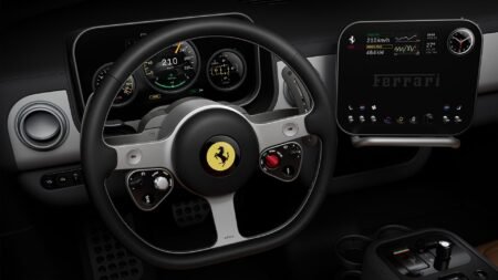 Ferrari Luce interior cockpit: Black leather-wrapped steering wheel with iconic prancing horse emblem, integrated controls, and red engine start button. Behind it, a multi-gauge digital instrument cluster displays speed (210 km/h), range, and vehicle data. To the right, a large central touchscreen shows performance metrics (464 kW power) and the Ferrari logo, with climate and connectivity controls below.