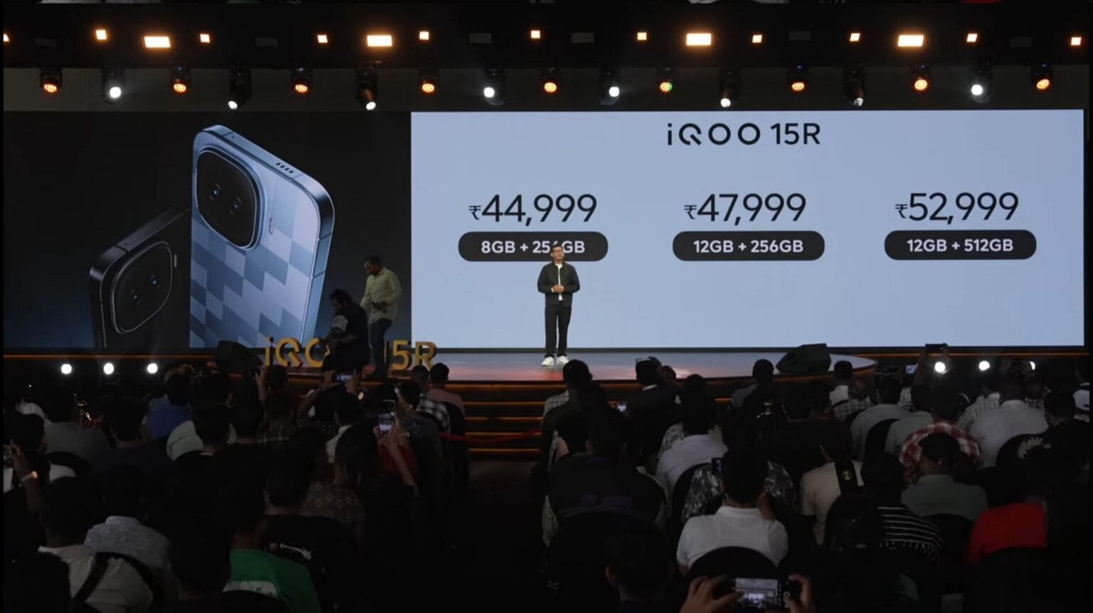 A presenter stands on stage during the iQOO 15R launch event, with a large screen behind showing the phone design and official prices in India: ₹44,999 for 8GB+256GB, ₹47,999 for 12GB+256GB, and ₹52,999 for 12GB+512GB, while an audience watches and records the presentation.