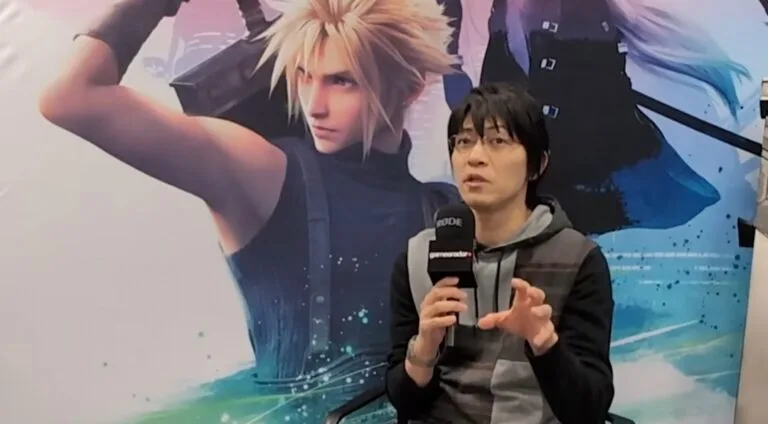 Naoki Hamaguchi, the director of the Final Fantasy VII Remake series, speaks into a "RODE gamereadar" microphone. He is wearing glasses and a gray and black hooded sweatshirt, gesturing with his hands. Behind him is a large promotional image of Cloud Strife from Final Fantasy VII Remake, with his signature spiky blonde hair and Buster Sword.