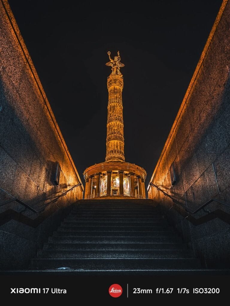 A low-angle shot looking up a wide staircase towards the brightly lit Golden Victoria statue atop the Berlin Victory Column at night. The stairs are dark, contrasting with the warm orange glow on the walls and the monument. Text at the bottom indicates "XIAOMI 17 Ultra," "Leica," and camera settings "23mm f/1.67 1/7s ISO3200.