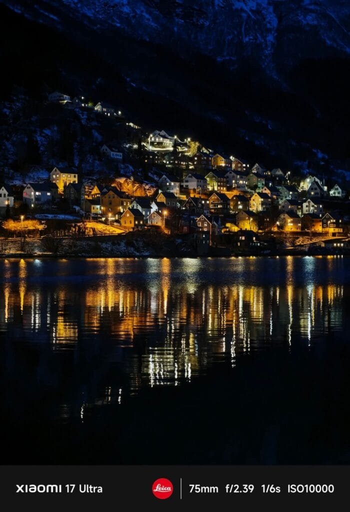 A vibrant night scene of a village nestled on a hillside, illuminated by warm lights that reflect beautifully on the dark, rippling water of a fjord or lake below. Snow-dusted mountains are visible in the background. Text at the bottom indicates "XIAOMI 17 Ultra," "Leica," and camera settings "75mm f/2.39 1/6s ISO10000.