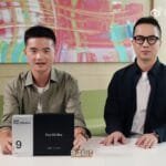 Two people at a table; left holds light gray Find X9s Pro and wider black Find X9 Ultra boxes. Combined width spans nearly outstretched hands, heights match, Ultra box slightly thicker.