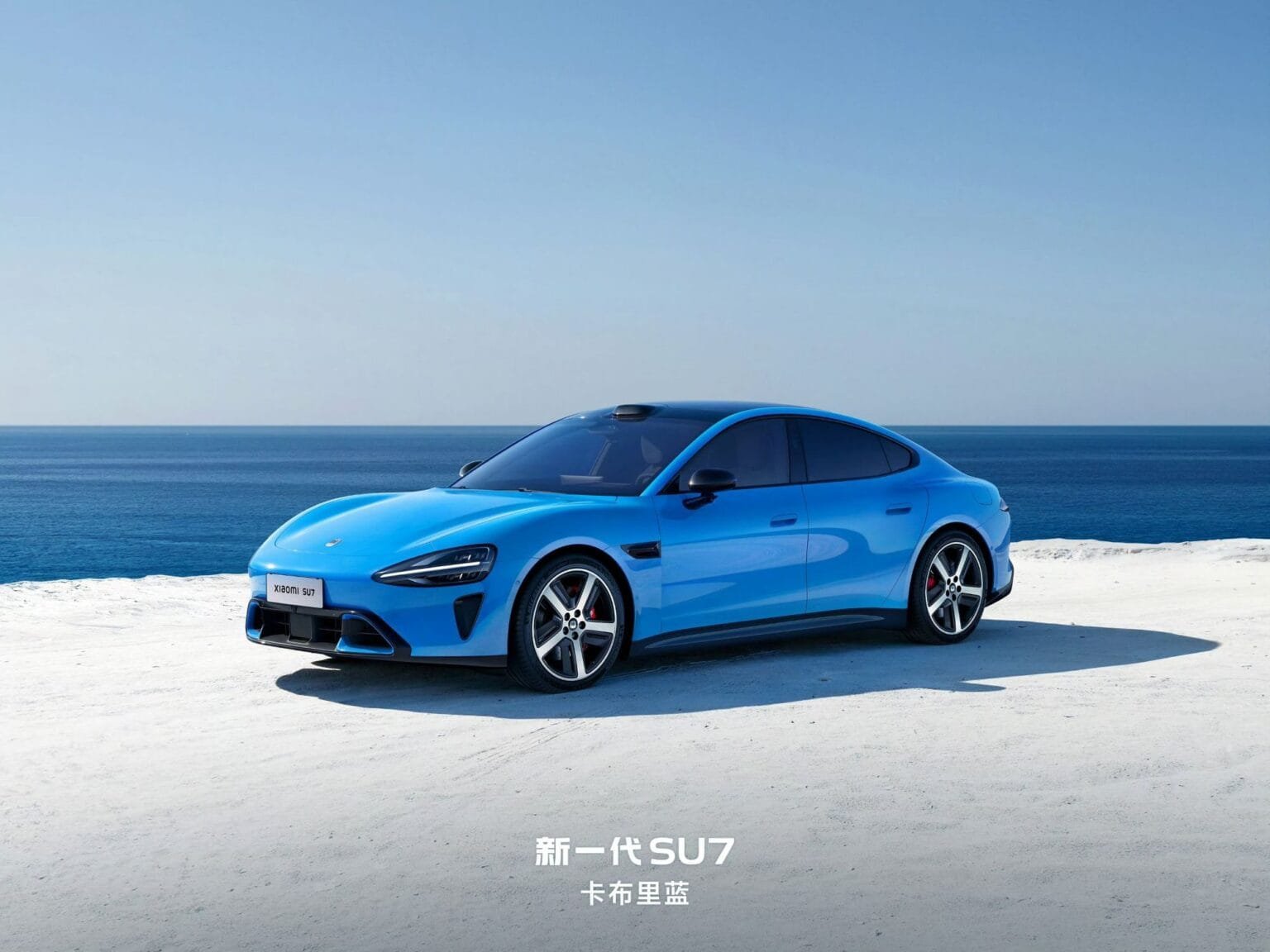 A blue Xiaomi SU7 electric car parked on a sandy beach with the ocean in the background.