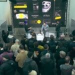 Overhead view of a product launch event of Nothing Phone 4a and Phone 4a Pro with a crowd gathered in front of a stage, where two speakers are presenting in front of a large screen displaying mobile interface graphics.