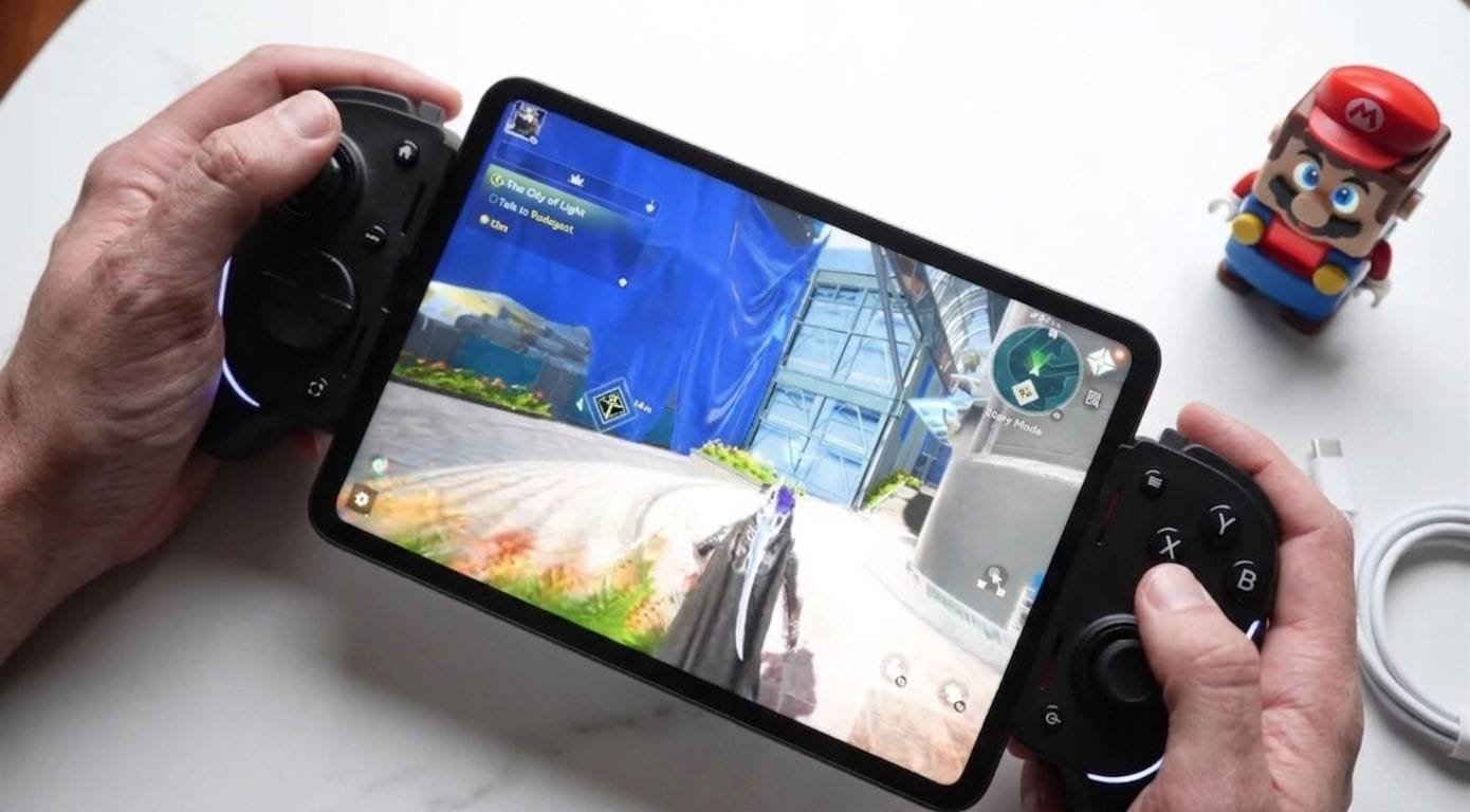 Xiaomi Pad mini tablet held by hands with attached game controllers, displaying a 3D open-world game interface on its screen. A small Mario figurine and charging cable are visible on the white surface beside it.