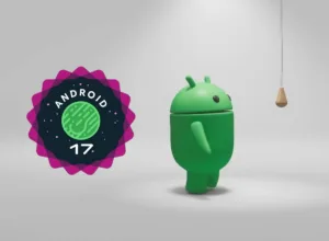 Green Android mascot standing next to the Android 17 badge, looking up at a small hanging wooden weight in a gray studio.