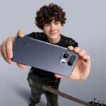 Young person holding a sleek, dark blue realme smartphone from the back, showcasing its modern camera module design; they stand against a plain light gray background, with a black electric bass guitar positioned at their side.