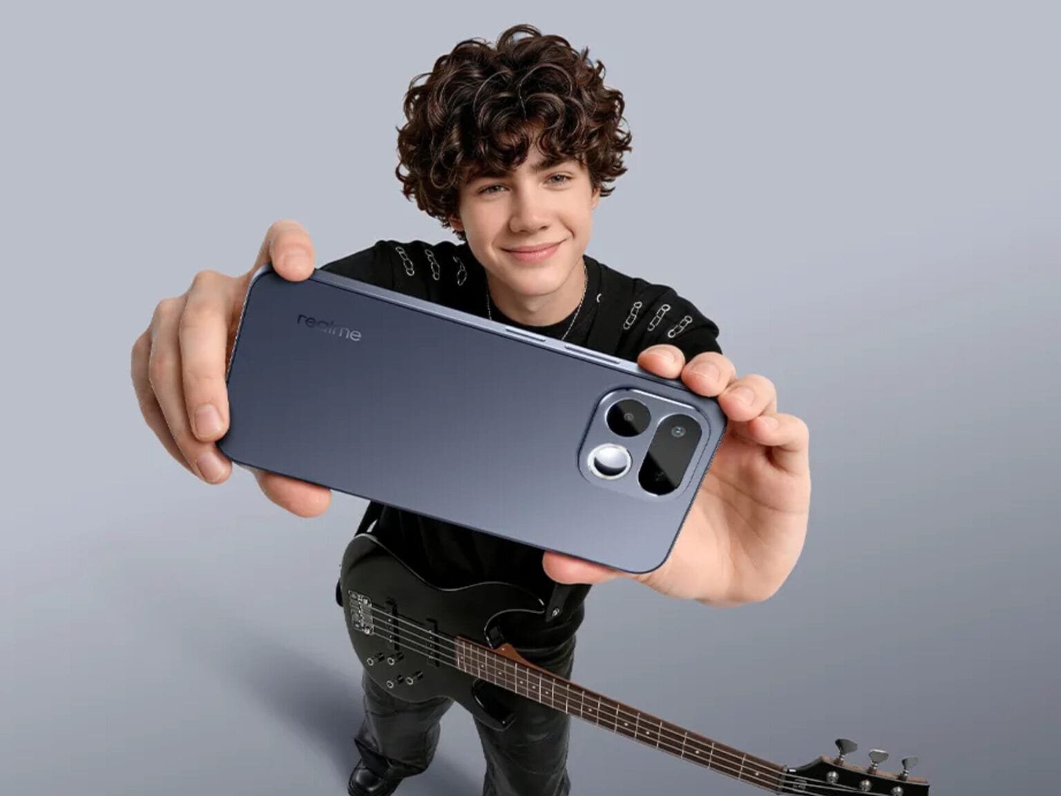 Young person holding a sleek, dark blue realme smartphone from the back, showcasing its modern camera module design; they stand against a plain light gray background, with a black electric bass guitar positioned at their side.