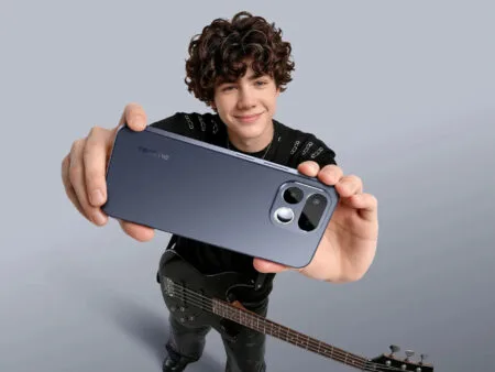 Young person holding a sleek, dark blue realme smartphone from the back, showcasing its modern camera module design; they stand against a plain light gray background, with a black electric bass guitar positioned at their side.