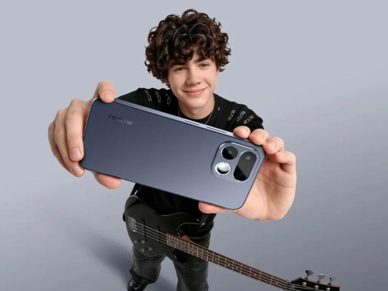 Young person holding a sleek, dark blue realme smartphone from the back, showcasing its modern camera module design; they stand against a plain light gray background, with a black electric bass guitar positioned at their side.