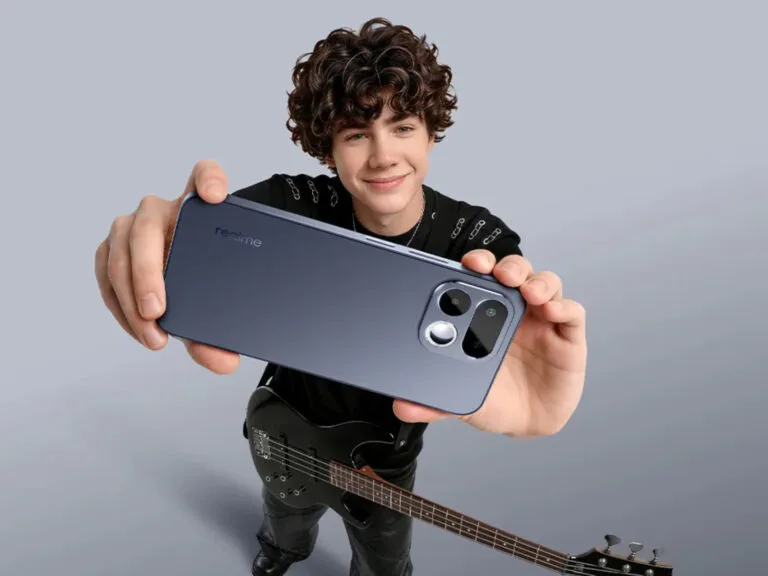 Young person holding a sleek, dark blue realme smartphone from the back, showcasing its modern camera module design; they stand against a plain light gray background, with a black electric bass guitar positioned at their side.