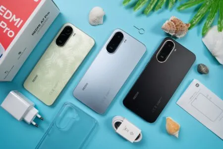Flat-lay shot of three Redmi A7 Pro smartphones in light green, light blue, and black colorways, displayed alongside their retail box, 15W charger, USB cable, clear protective case, SIM tool, and user manual, arranged on a bright blue surface decorated with seashells and green leaves.