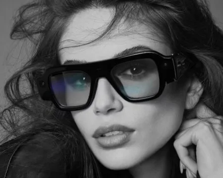 Snap SPECS AR glasses shown in glossy black frame design, worn in black‑and‑white studio shot; visible camera module on side arm, bold angular silhouette, reflective lenses; modern wearable tech with stylish eyewear form factor.