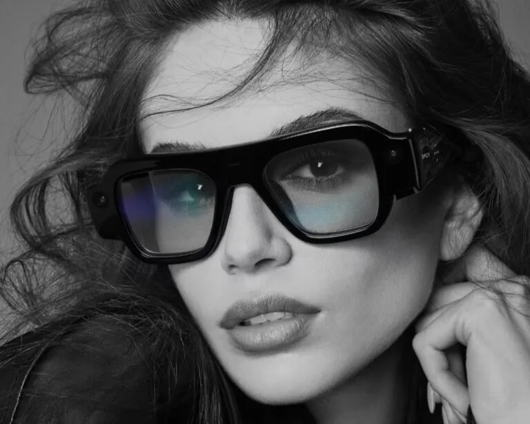 Snap SPECS AR glasses shown in glossy black frame design, worn in black‑and‑white studio shot; visible camera module on side arm, bold angular silhouette, reflective lenses; modern wearable tech with stylish eyewear form factor.