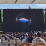 Outdoor event at Apple Park with a large screen displaying a colorful swirling graphic, attendees seated facing the stage