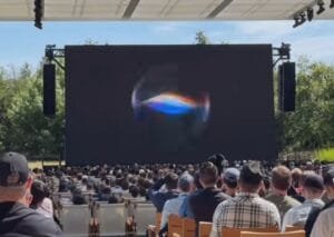 Outdoor event at Apple Park with a large screen displaying a colorful swirling graphic, attendees seated facing the stage