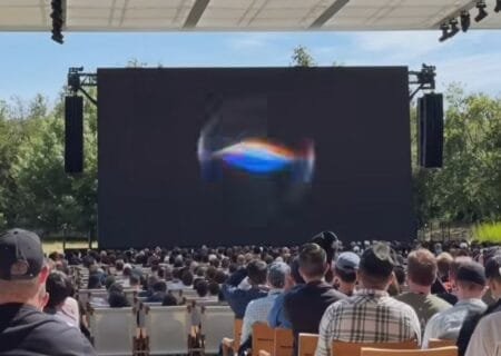 Outdoor event at Apple Park with a large screen displaying a colorful swirling graphic, attendees seated facing the stage