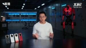 Screenshot from the replay of the HONOR X80 Pro Max "X-Journey" new product launch event; a presenter in a white t-shirt speaks at a table displaying four color variants of the HONOR X80 Pro Max smartphone, with a red-and-black HONOR-branded humanoid robot standing beside him in a futuristic tech studio setting.