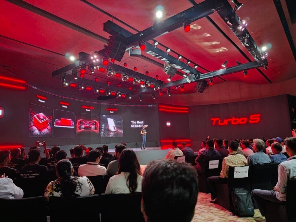 Redmi Turbo 5 Launched in India; Price Starts at ₹37,999 1 Redmi Turbo 5 official launch event in India stage view with presenter product showcase screens red lighting and invited guests in attendance.