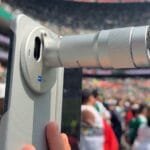Vivo X Fold 6 Camera Specifications Revealed 4 Vivo X Fold 6 foldable smartphone with extended telephoto lens accessory, ZEISS certified camera system, light textured rear panel, captured at stadium event for long distance shooting.