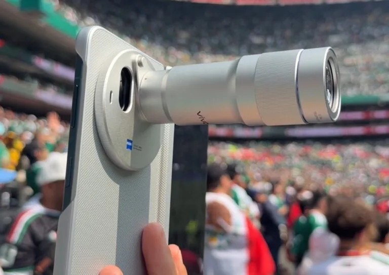 Vivo X Fold 6 Camera Specifications Revealed Vivo X Fold 6 foldable smartphone with extended telephoto lens accessory, ZEISS certified camera system, light textured rear panel, captured at stadium event for long distance shooting.