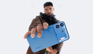 Product shot of the blue Nothing Phone (4b) held out toward the camera, showing its smooth matte back panel, distinct rectangular camera module with dual lenses, and the new compact Glyphs Bar light element. A person in a brown leather jacket holds the phone against a plain light background.