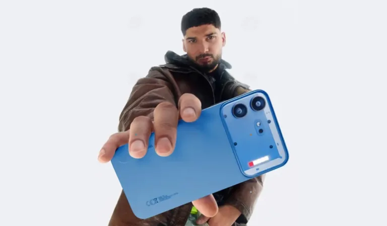 Product shot of the blue Nothing Phone (4b) held out toward the camera, showing its smooth matte back panel, distinct rectangular camera module with dual lenses, and the new compact Glyphs Bar light element. A person in a brown leather jacket holds the phone against a plain light background.