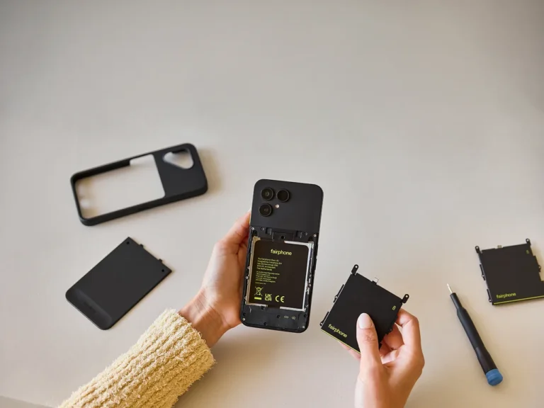 Hands holding the Fairphone Gen 6+ with the back cover removed. The phone shows its modular internal design with a removable battery and replaceable parts. A repair tool sits nearby to demonstrate the device's easy repairability.